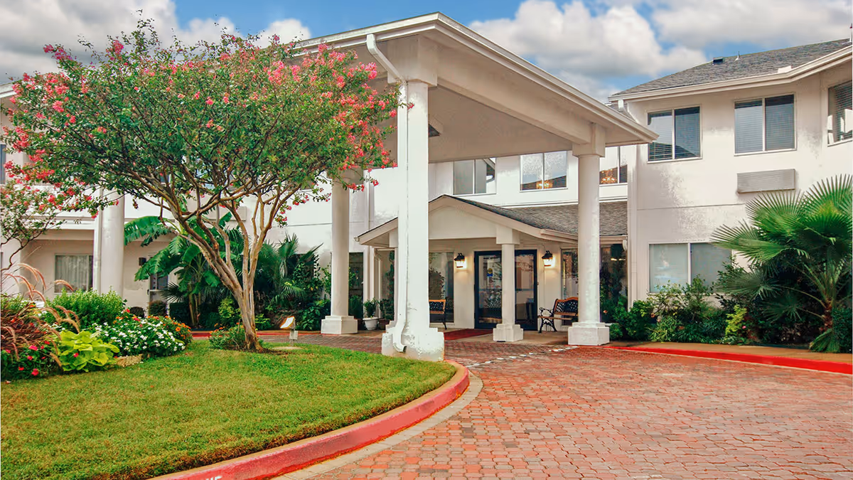Entrance of a senior living facility with a covered driveway, surrounded by well-maintained landscaping including a flowering tree, bushes, and green grass under a partly cloudy sky.
