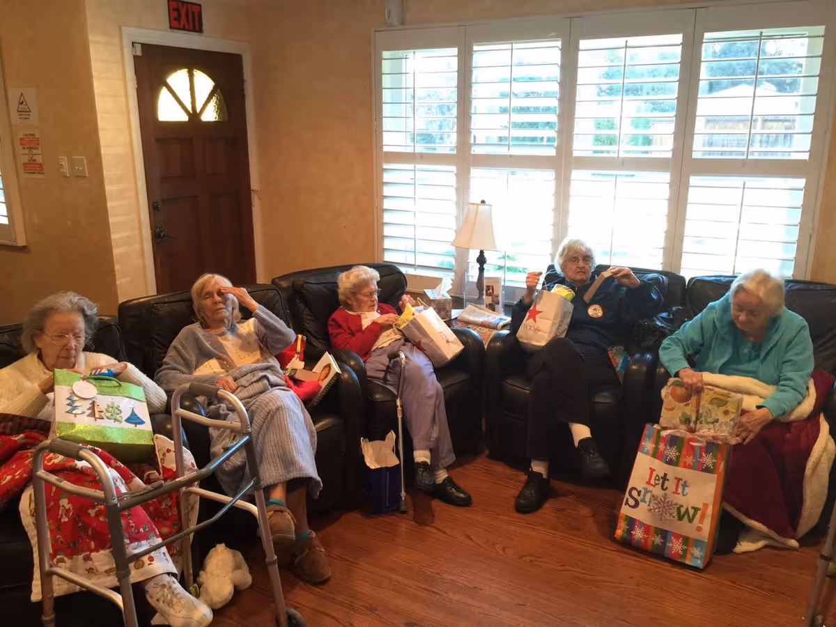 Five elderly women sitting on black leather chairs in a living room with wooden floors and large windows with white shutters. They are holding holiday gift bags and appear to be opening presents. There is a walker in front of one woman and a table lamp on a side table near the window.