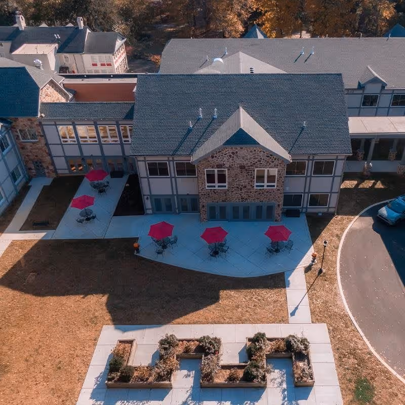 Aerial view of a senior living facility building with a stone and siding exterior. The building has a patio area with several tables and red umbrellas. Surrounding the patio is a lawn with a garden area featuring raised flower beds. A curved driveway is visible on the right side of the image.