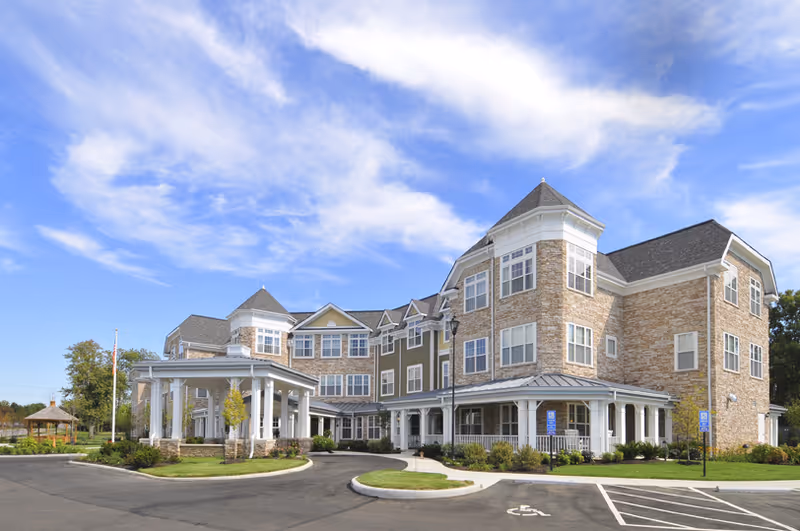 Exterior view of a large, multi-story senior living facility building with stone and siding facade, multiple windows, a covered entrance, and a circular driveway under a partly cloudy blue sky.