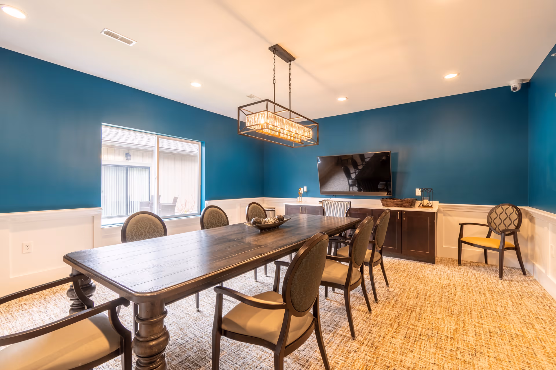A well-lit meeting or dining room with a long wooden table surrounded by eight upholstered chairs. The walls are painted teal with white wainscoting, and there is a large flat-screen TV mounted on the wall. A modern rectangular chandelier hangs above the table, and a window lets in natural light. The floor is carpeted with a textured pattern.