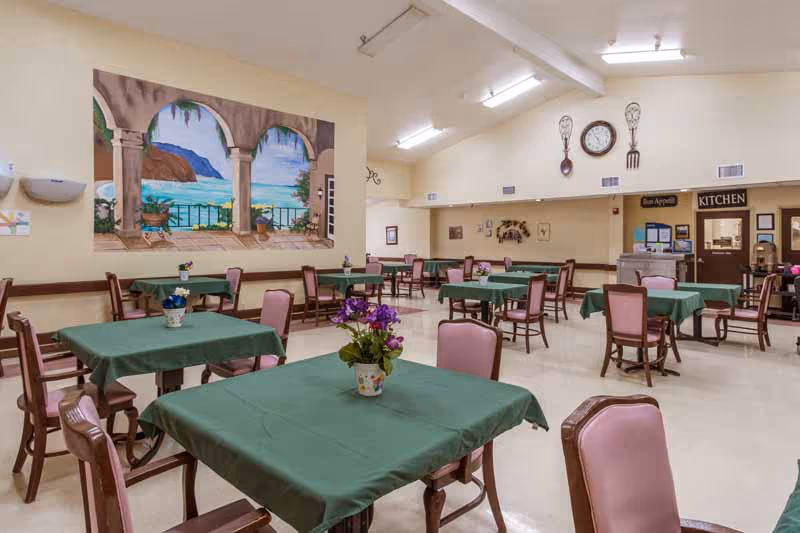 Spacious dining room with multiple tables covered in green tablecloths and pink chairs, a large seaside mural on the wall and a service window labeled "KITCHEN".