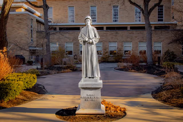A statue of a woman in a long robe holding a book stands on a pedestal in front of a brick building with multiple windows. The statue is surrounded by a circular paved area with some bushes and trees nearby.