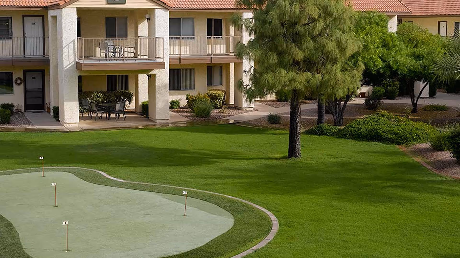 Outdoor view of Atria Chandler Villas showing a putting green with four small flags, surrounded by well-maintained green grass, trees, and shrubs. In the background, there is a two-story building with balconies and patio seating areas.