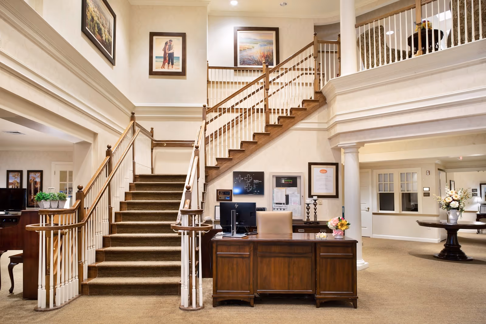 Interior view of a senior living facility lobby with a wooden reception desk, a staircase with wooden handrails and white balusters, framed artwork on the walls, and a round table with a floral arrangement. The space is warmly lit and carpeted.