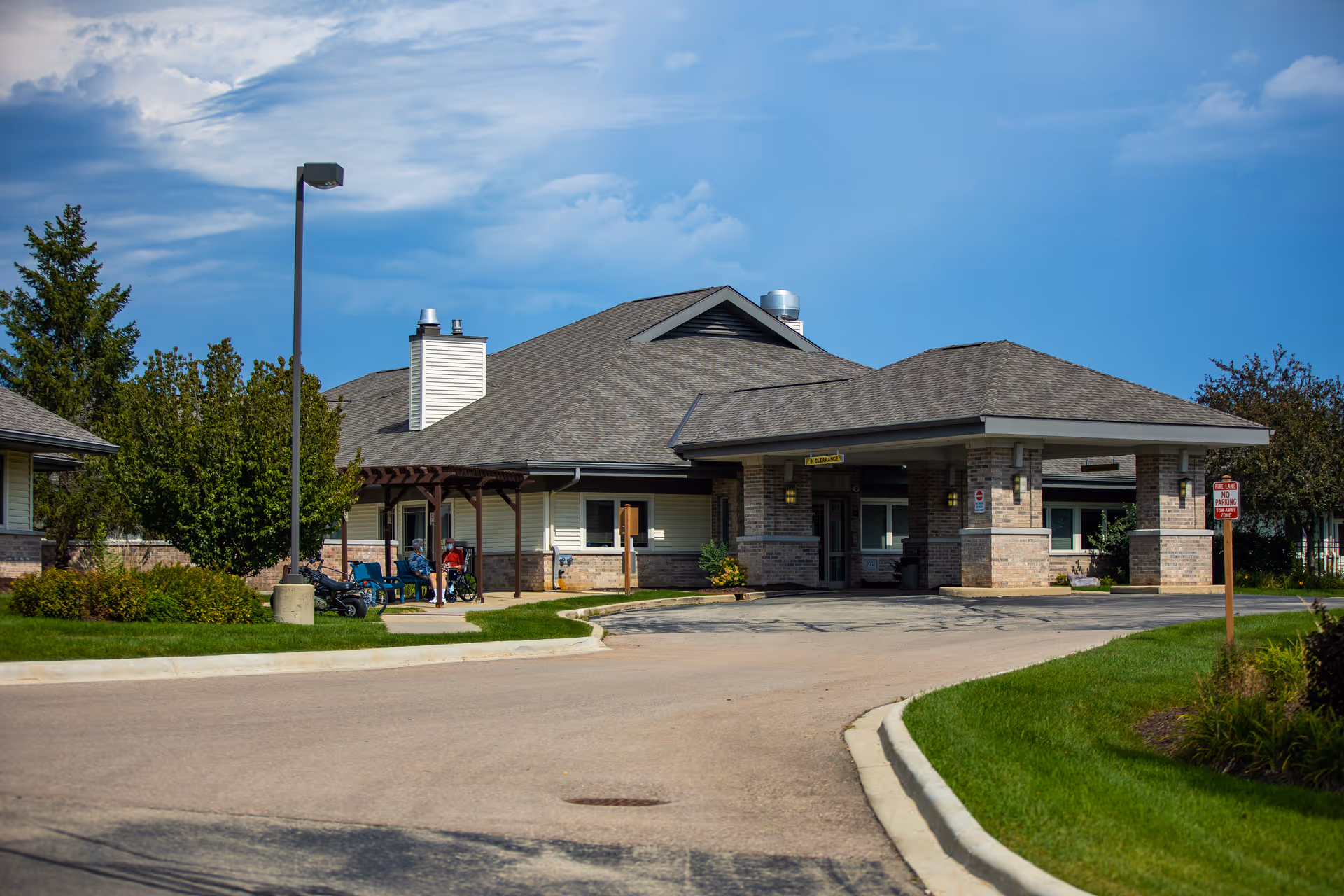 Exterior view of St. Mary's Care Center showing the main entrance with a covered drop-off area. The building has a pitched roof and brick and siding exterior. There are a few people sitting on benches near the entrance, and the surrounding area has green grass, trees, and a clear blue sky.