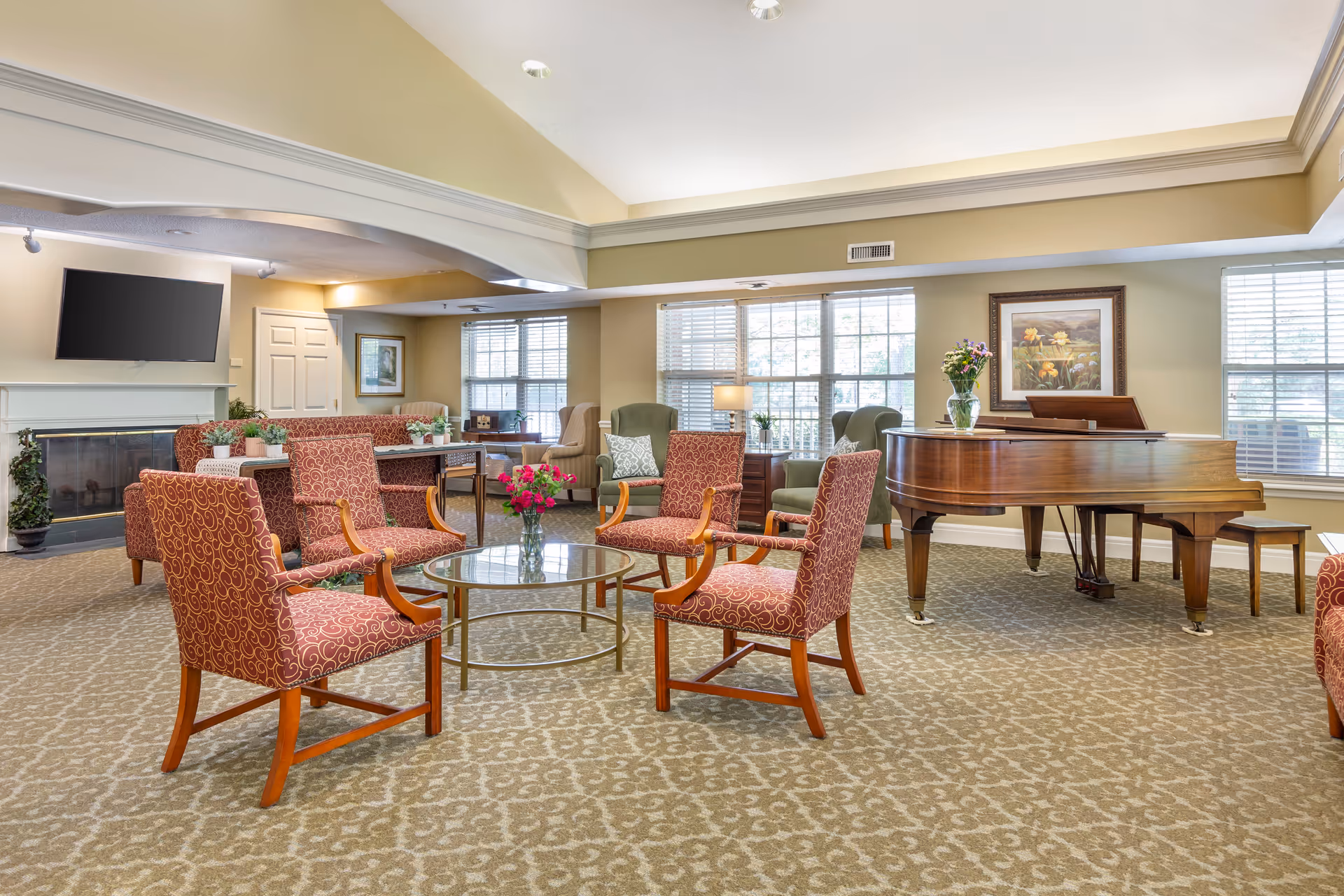Bright common living room with upholstered chairs arranged around a glass coffee table, a grand piano, fireplace, and large windows.