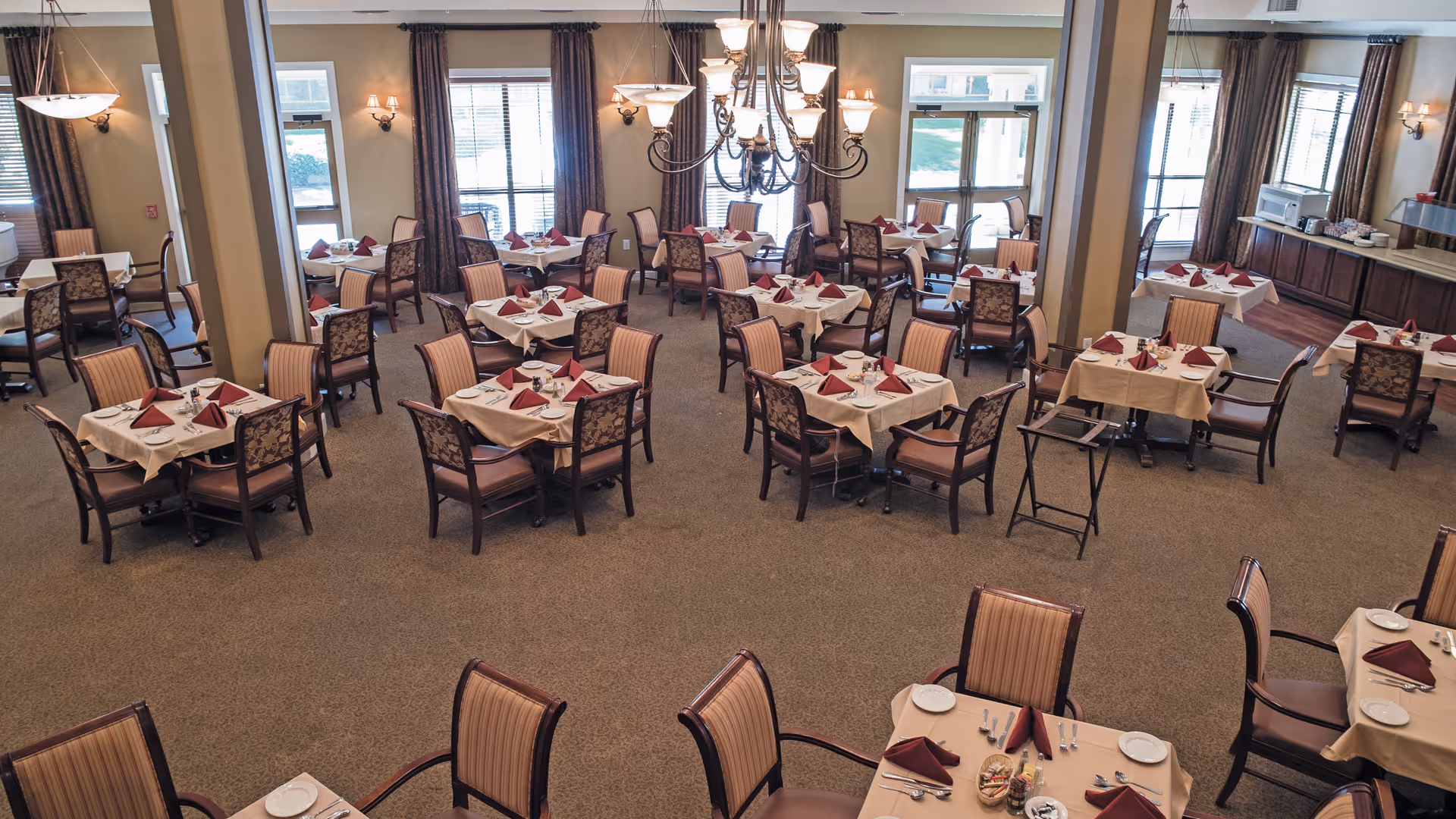 Spacious dining room with multiple tables set with white linens, folded red napkins, and chairs under chandeliers.