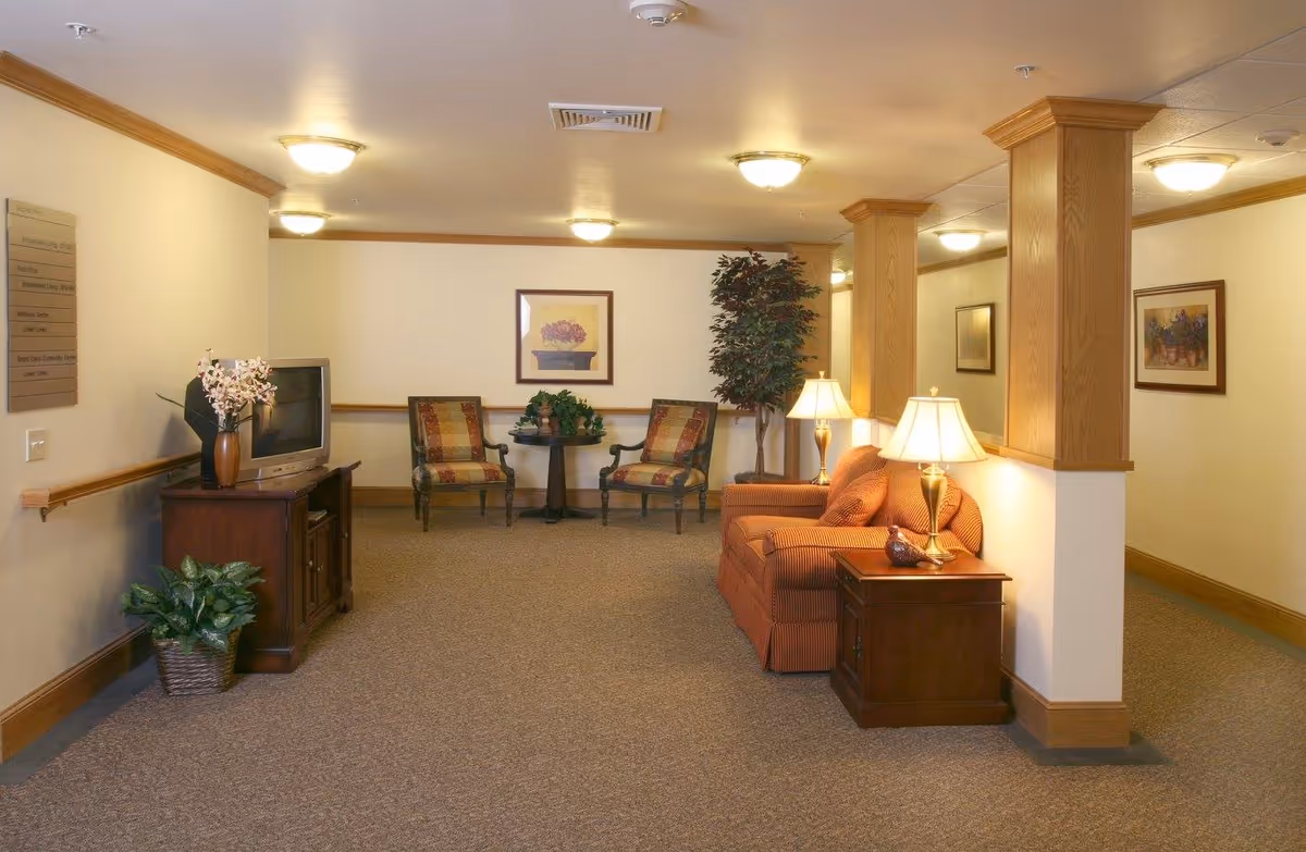 A cozy common area with a striped orange sofa, two patterned armchairs, a small round table with plants, a wooden TV stand with a television and a vase of flowers, and several lamps providing warm lighting. The room has beige walls, carpeted floor, wooden trim, and decorative columns.
