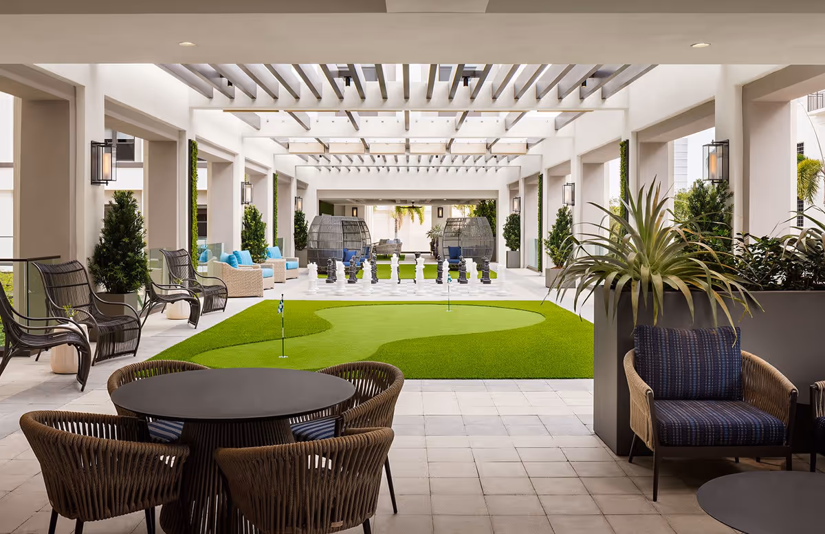 Covered outdoor seating area with wicker chairs and tables surrounding a putting green and a large chessboard with oversized chess pieces. The space is decorated with potted plants and has a modern pergola overhead.