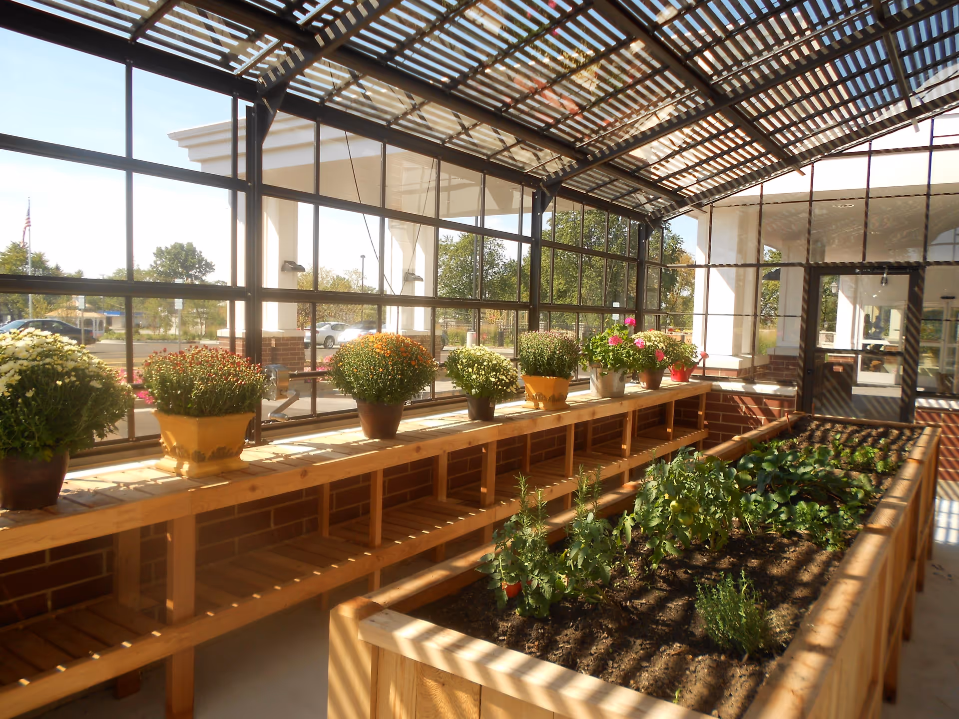 Indoor garden room with a wooden raised garden bed filled with various plants and herbs. Along the window ledge, there are several potted flowering plants. The room has large glass windows and a slatted roof allowing sunlight to enter.