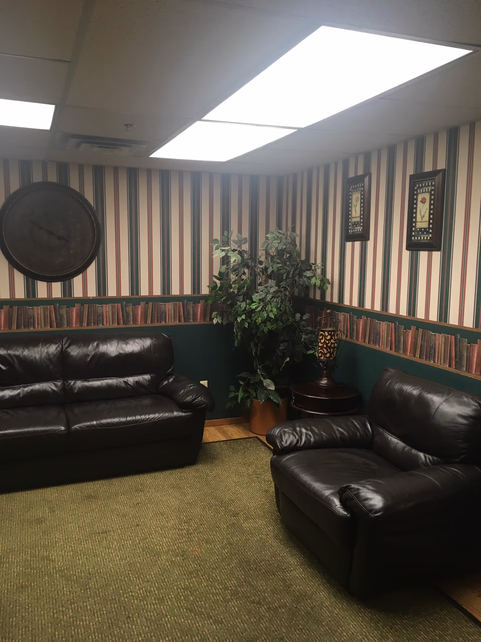 Small lounge with dark leather sofas and an armchair, striped wallpaper, a potted plant and overhead fluorescent lights.