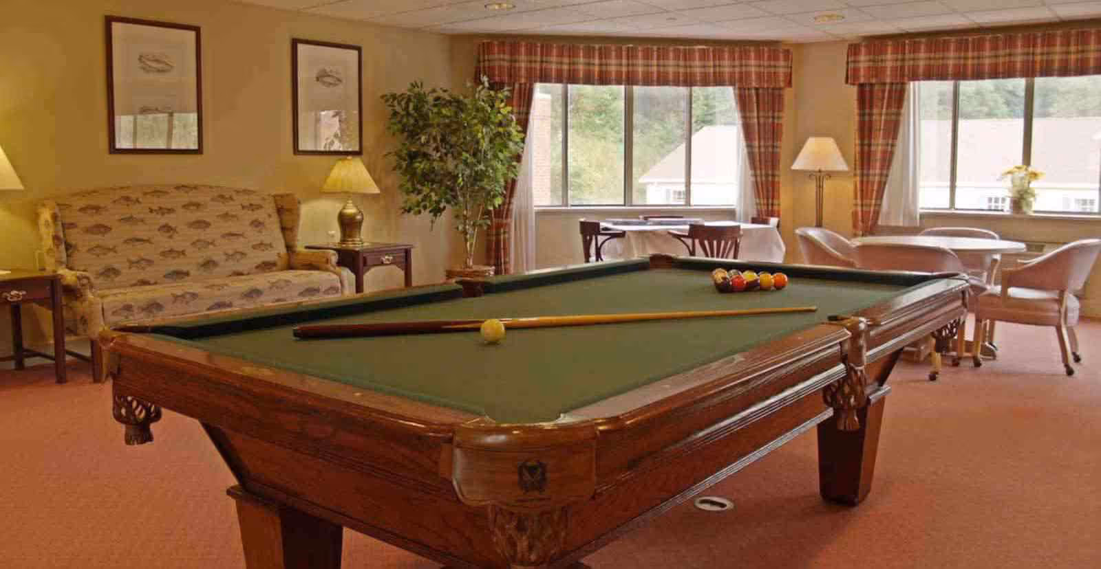 A cozy common room featuring a pool table with a cue stick and balls arranged on it. Behind the pool table, there is a patterned sofa with two framed pictures above it, a potted plant, and two lamps on side tables. The room has large windows with plaid curtains letting in natural light, and there are tables and chairs near the windows.