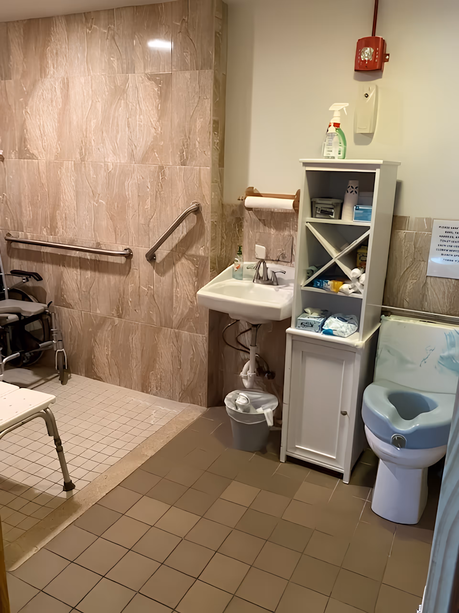 Accessible bathroom with beige tiled walls and floor, featuring a shower area with grab bars and a shower chair. There is a small white sink with a soap dispenser, a white storage cabinet with various supplies, and a toilet with a raised seat and armrests. A red fire alarm is mounted on the wall above the cabinet.