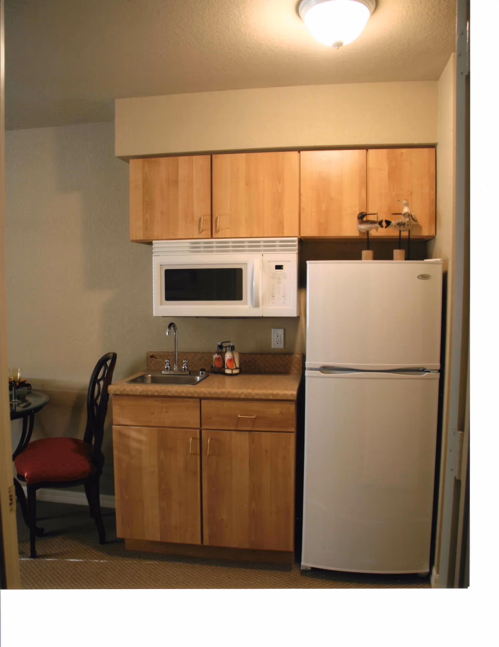 A small kitchenette with light wood cabinets, a white microwave mounted above a countertop with a sink, and a white refrigerator. There are two decorative bird figurines on top of the refrigerator. To the left, a partial view of a glass-top table with a red cushioned chair is visible.