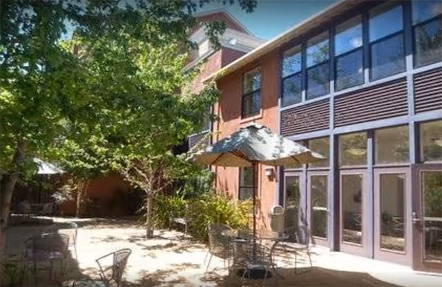Sunlit courtyard with patio tables and umbrellas next to a two-story building with large windows.