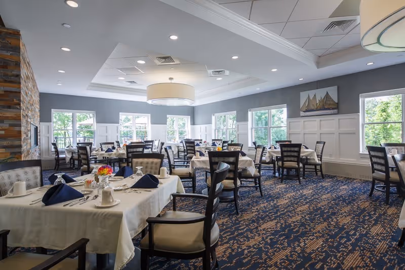 A bright and spacious dining room with multiple tables covered in white tablecloths, each set with plates, cups, silverware, and navy blue napkins. The room has large windows letting in natural light, a patterned blue carpet, and a modern ceiling light fixture. There is a stone accent wall on the left side and a painting of sailboats on the far wall.