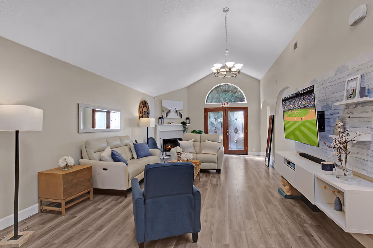 A bright and spacious living room in a senior living facility featuring beige walls, wood flooring, a beige leather sofa with blue cushions, a blue armchair, a wooden side table with a white flower arrangement, a fireplace, a wall-mounted flat-screen TV showing a baseball game, and a large arched window above double glass doors.