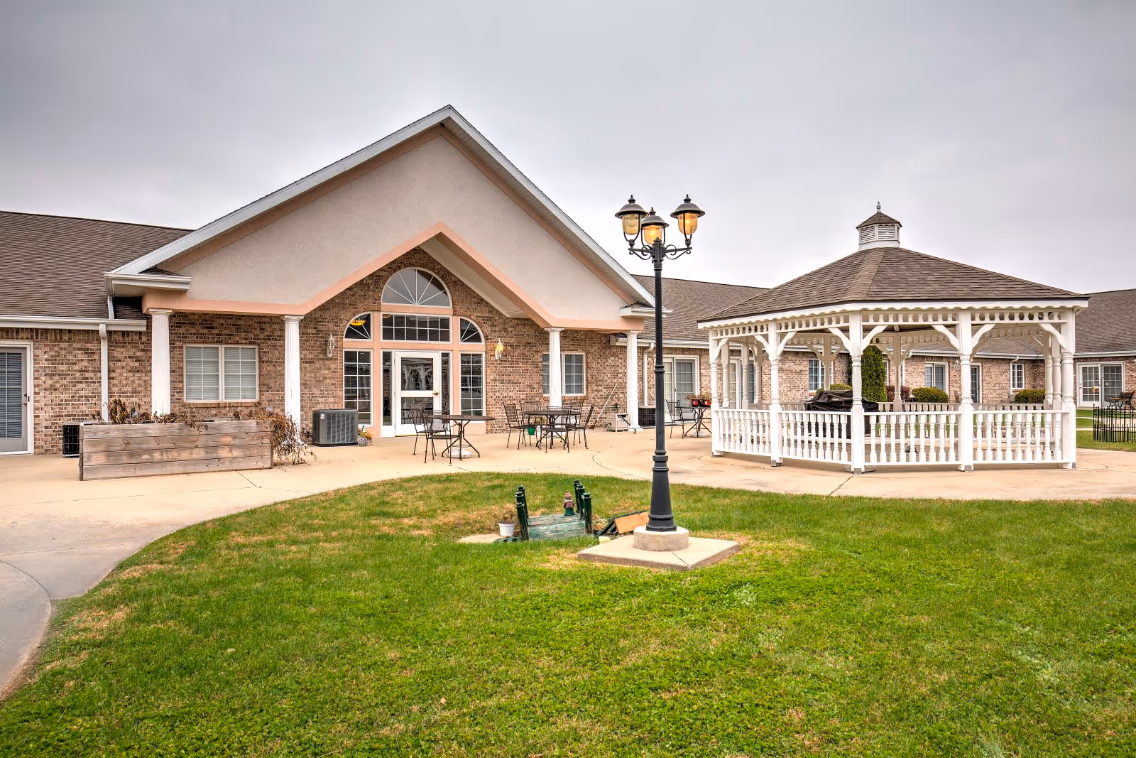 Outdoor view of Brookstone Estates of Charleston showing a brick building with a covered entrance, patio area with tables and chairs, a white gazebo, a black street lamp, and a green lawn in the foreground under an overcast sky.
