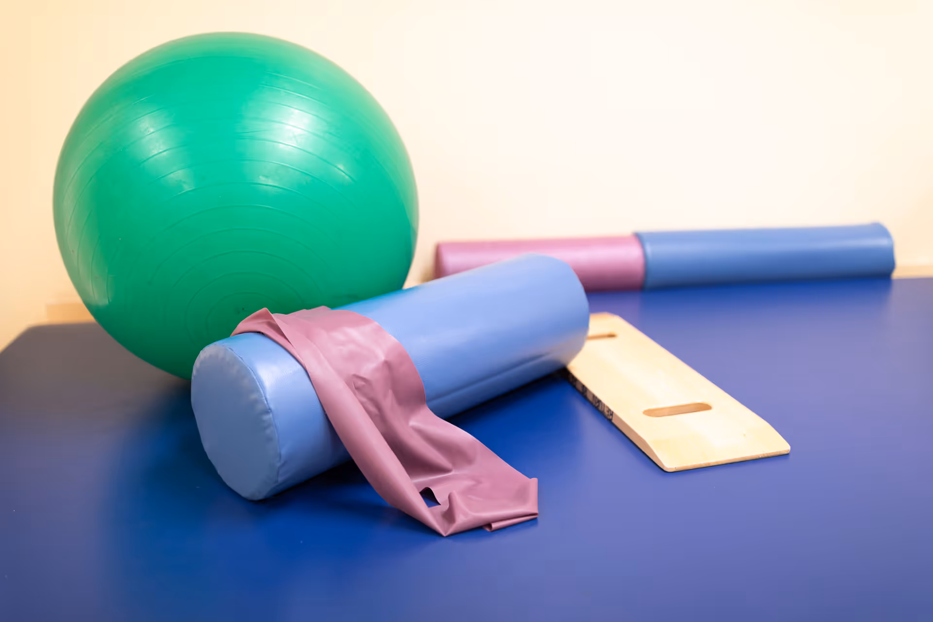 A green exercise ball, a blue cylindrical foam roller, a purple resistance band, and a wooden balance board placed on a blue exercise mat against a light-colored wall.
