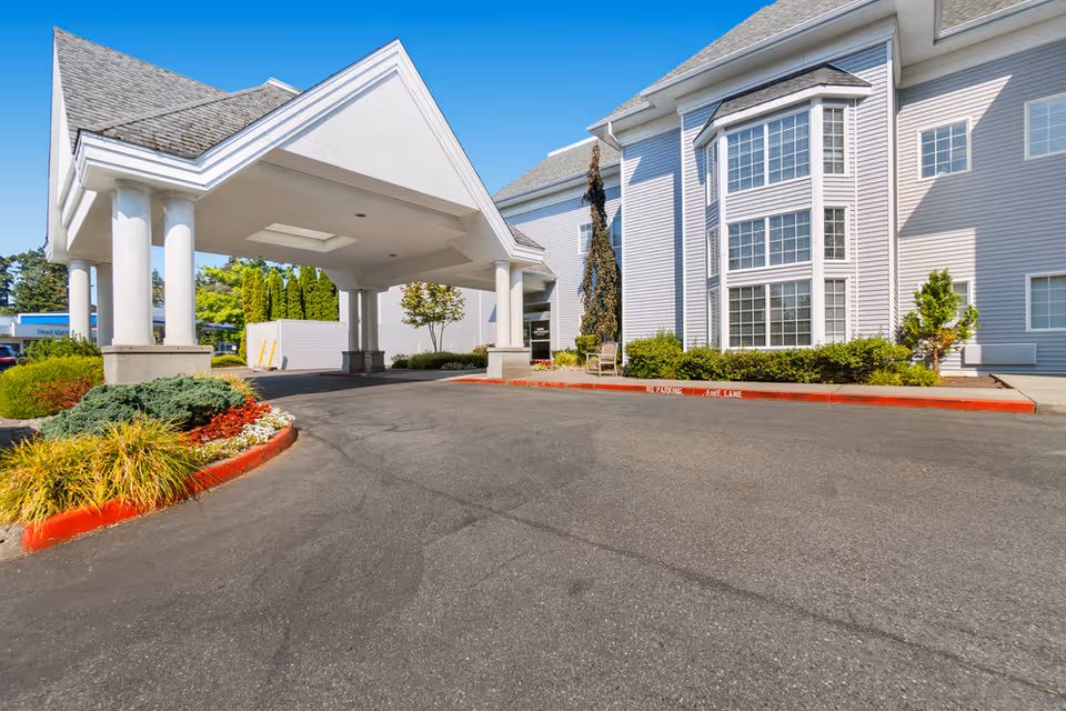 Covered porte-cochère and entrance drive of a light-gray multi-story senior living building with landscaping.