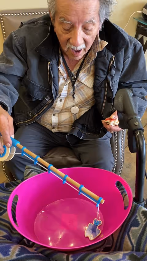 An elderly man sitting in a chair holding a toy fishing rod over a large pink plastic basin, appearing to play a fishing game indoors.
