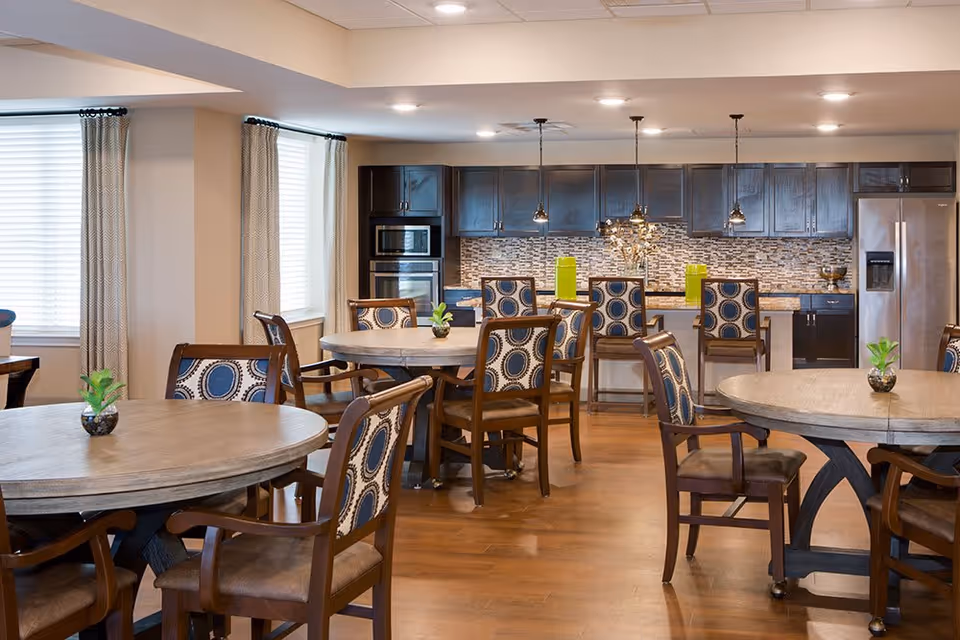 A spacious dining area with round wooden tables and cushioned chairs featuring a blue and white circular pattern. The room has large windows with light-colored curtains, wooden flooring, and a modern kitchen in the background with dark cabinets, stainless steel appliances, and pendant lighting above a counter with bar stools.