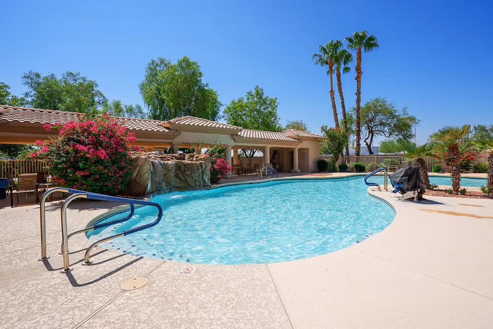 Outdoor swimming pool area at The Montecito Senior Living with clear blue water, surrounded by a concrete deck, tropical plants, palm trees, and a covered seating area with tables and chairs under a tiled roof. The sky is clear and blue.