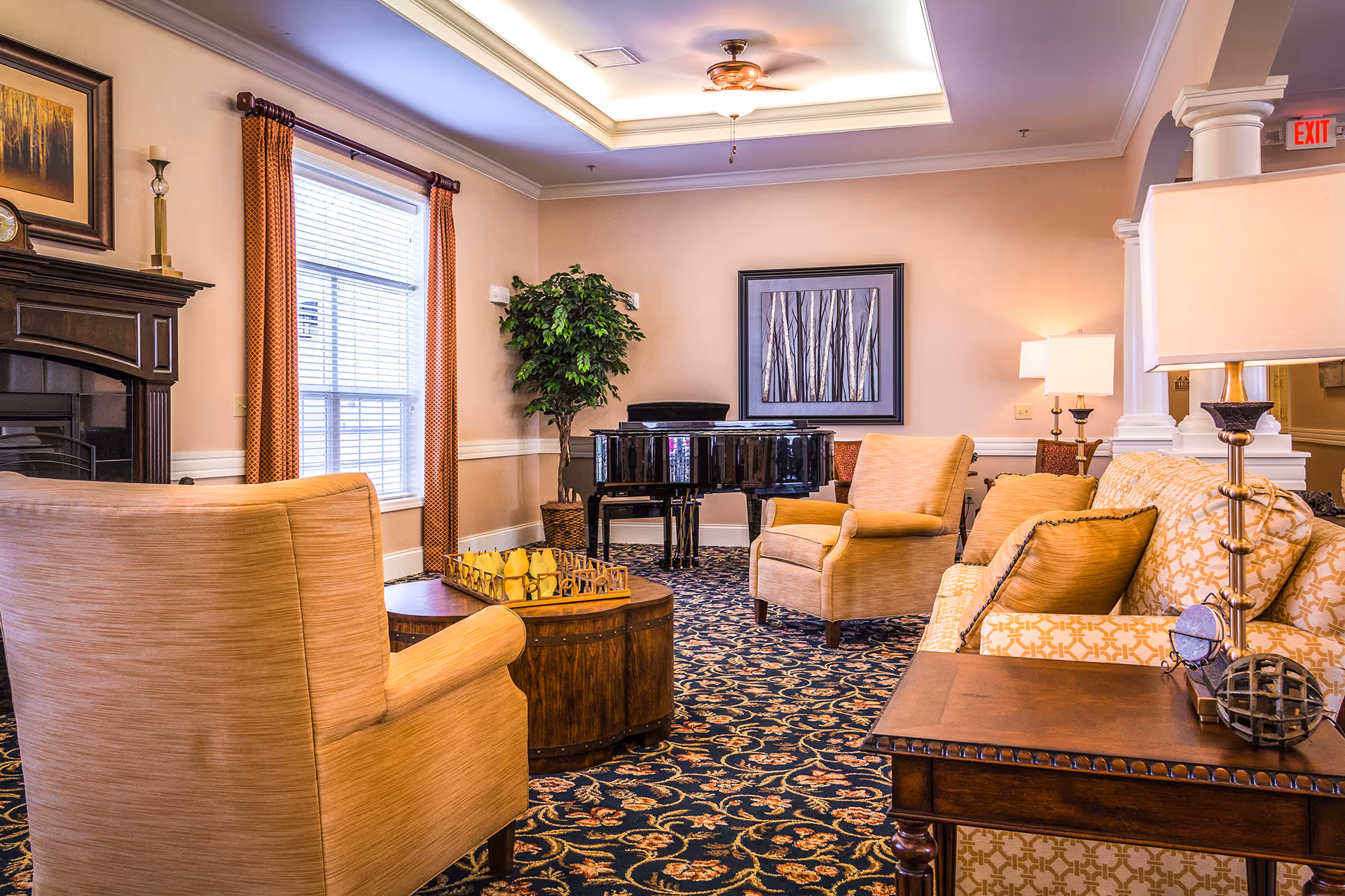 A cozy living room with beige armchairs and a patterned sofa arranged around a wooden coffee table. There is a black grand piano against the back wall, a large window with orange curtains, a potted plant, and framed artwork on the wall. The room features a decorative ceiling with a ceiling fan and warm lighting from table lamps.