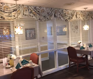 Small dining area with two set tables and chairs, pendant lights, and large interior windows with blinds and patterned valances.