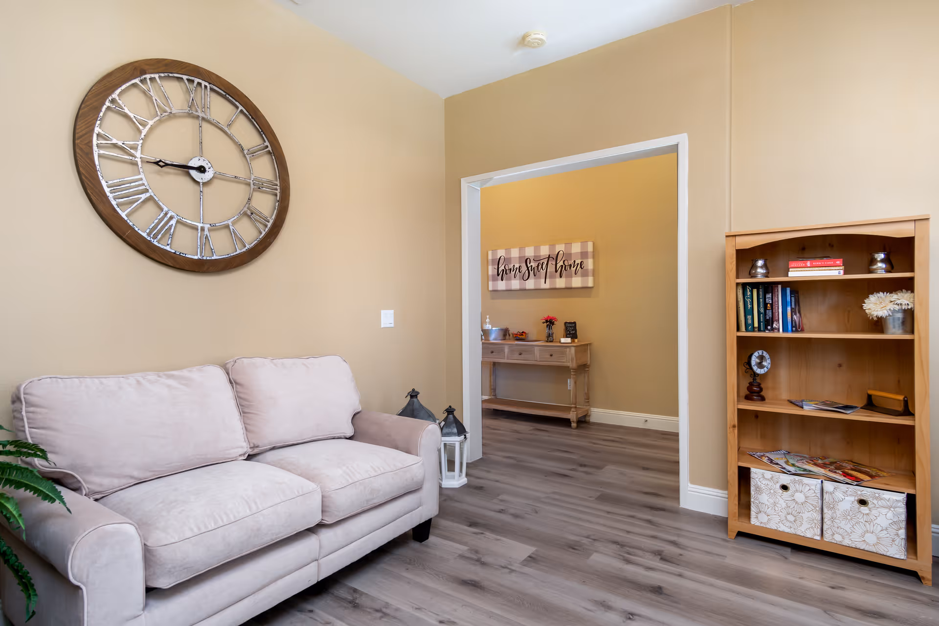 A cozy living room with a beige couch, a large decorative wall clock, a wooden bookshelf with books and decorative items, and a view into an adjacent room with a console table and a 'home sweet home' sign on the wall.