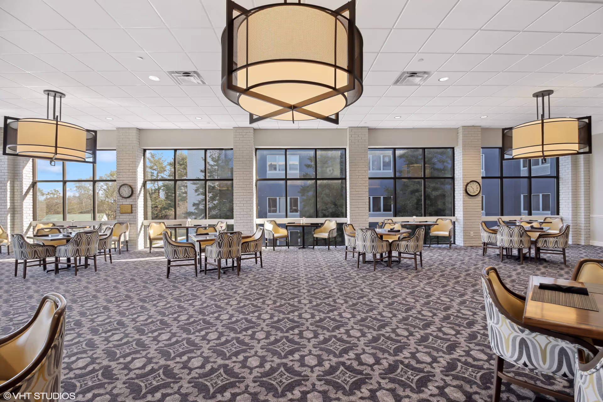 Spacious dining room with large windows allowing natural light to fill the space. The room features multiple tables with patterned chairs and beige cushioned seats. Large, modern pendant lights hang from the ceiling, and the floor is covered with a patterned carpet. Two clocks are mounted on white brick columns between the windows.