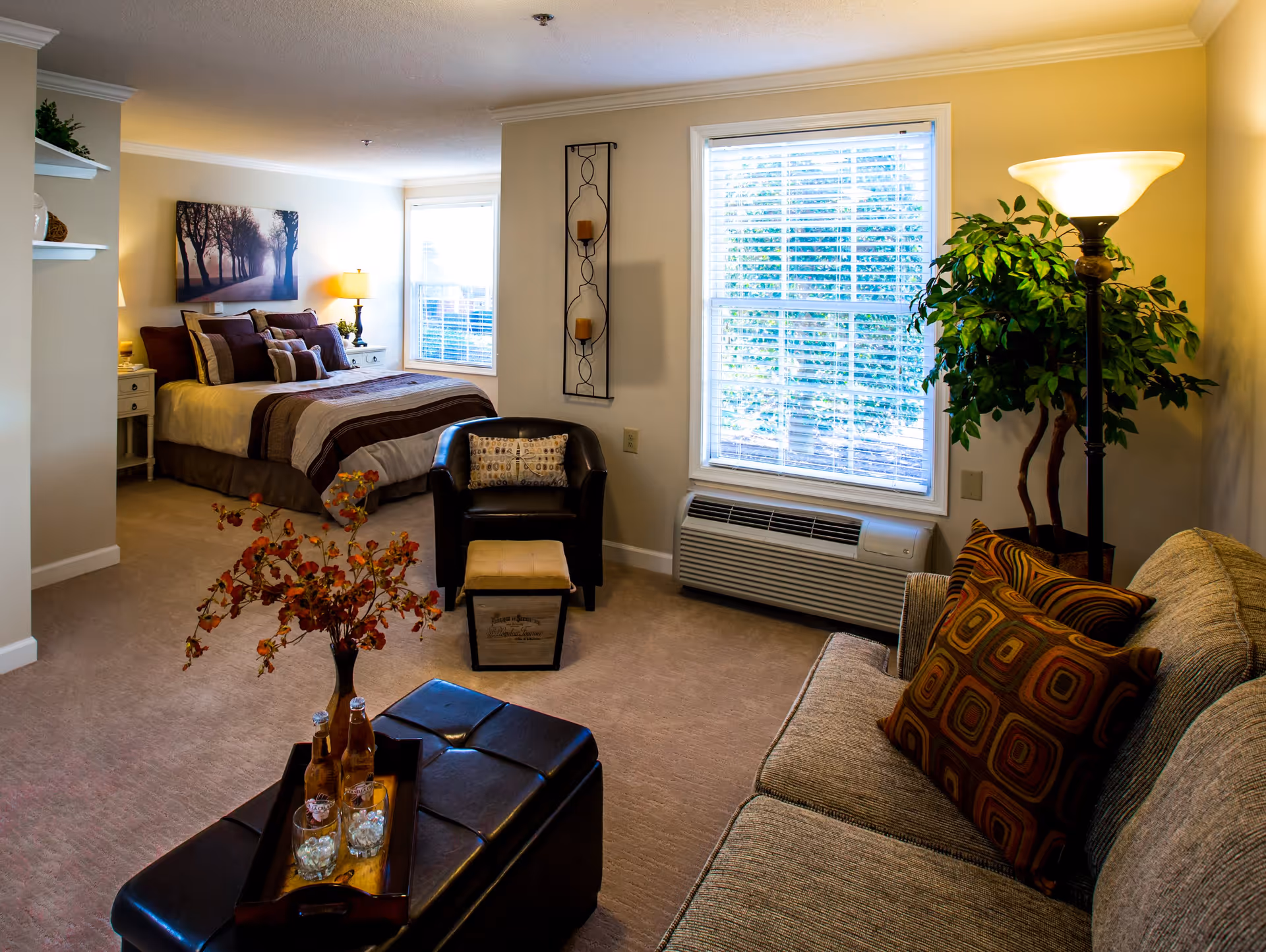 A cozy studio apartment interior with a bed in the background, a black armchair with a small ottoman, a beige sofa with patterned pillows, a tall floor lamp, a large window with blinds, and a black leather ottoman with a tray holding two bottles and two glasses. There is a decorative plant and wall art featuring trees on the wall behind the bed.