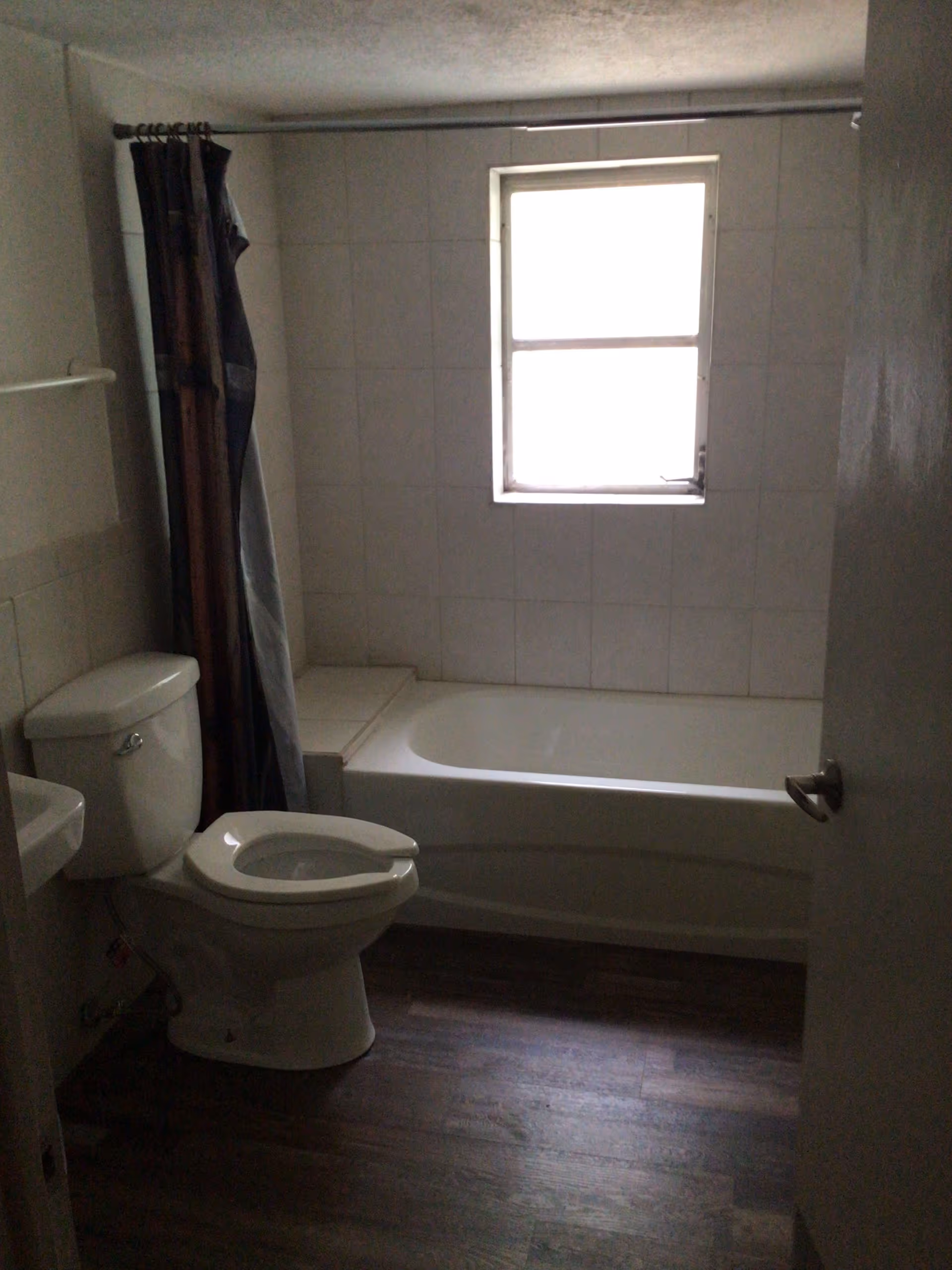 A bathroom with a white toilet, a bathtub with a shower curtain, a small window above the bathtub, and a door partially open. The floor appears to be wood or wood-like material.