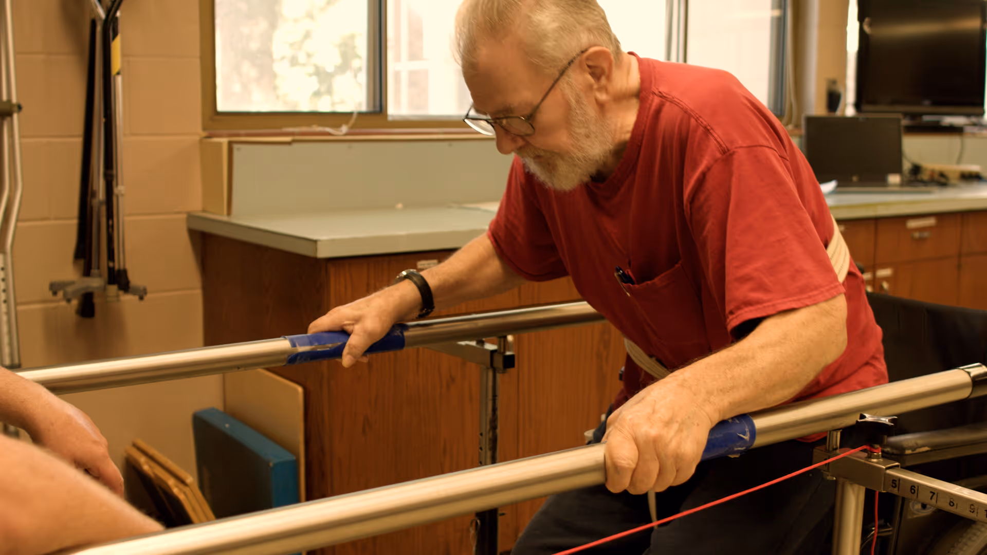 An elderly man wearing glasses and a red shirt is using parallel bars for physical therapy or rehabilitation in a room with wooden cabinets and a window in the background.