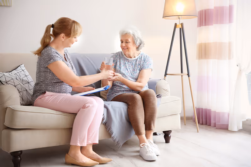 A young woman sitting on a beige couch handing a glass of water to an elderly woman who is also seated on the couch in a bright living room with a floor lamp and sheer curtains in the background.