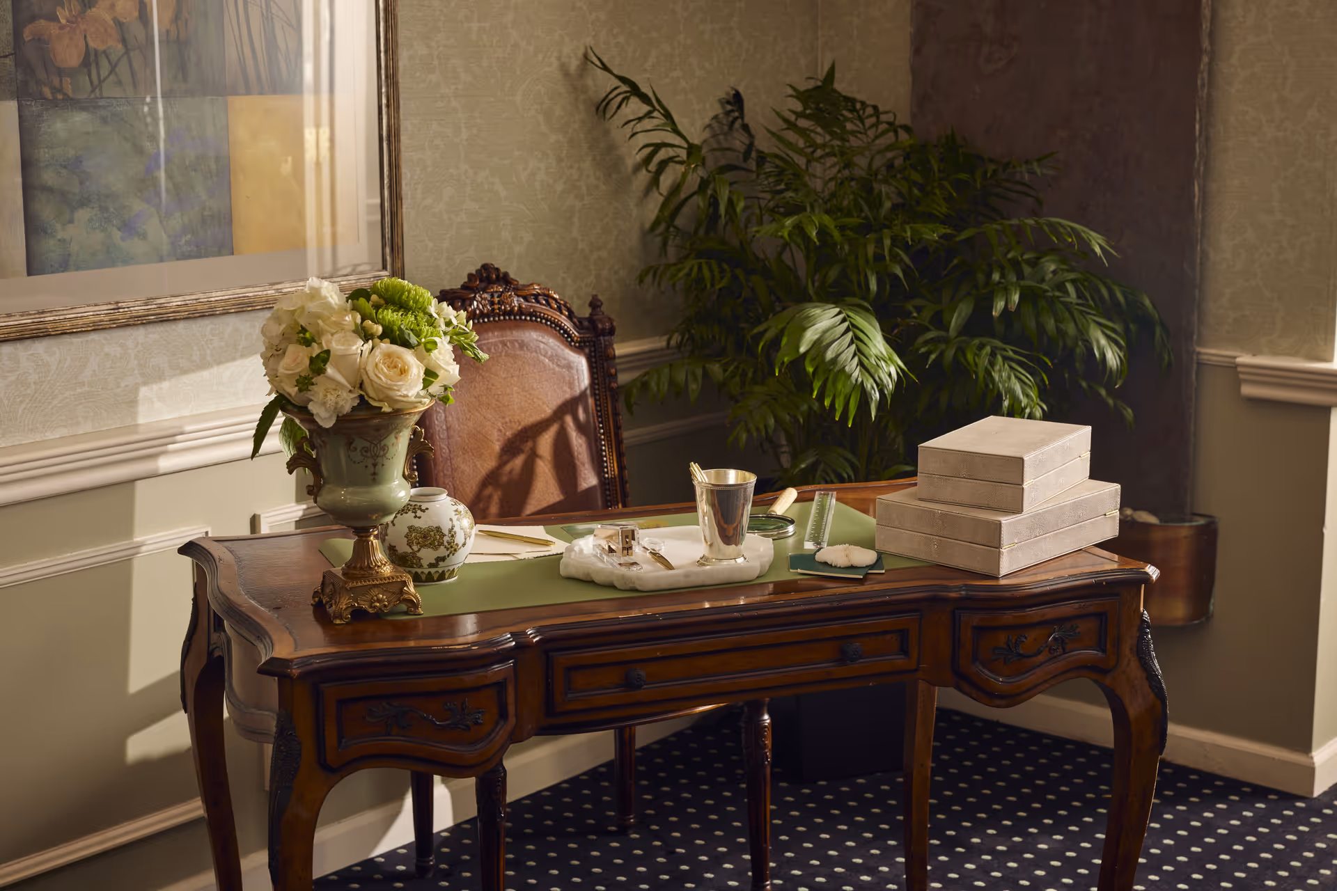 An elegant wooden desk with intricate carvings sits in a room with patterned wallpaper and a dark polka-dot carpet. On the desk are a green desk pad, a vase with white and green flowers, a small decorative jar, a silver cup, a magnifying glass, a ruler, some stationery, and three stacked beige boxes. Behind the desk is a vintage-style chair with a leather backrest, and a large green leafy plant is placed in the corner of the room. A framed abstract painting hangs on the wall to the left.