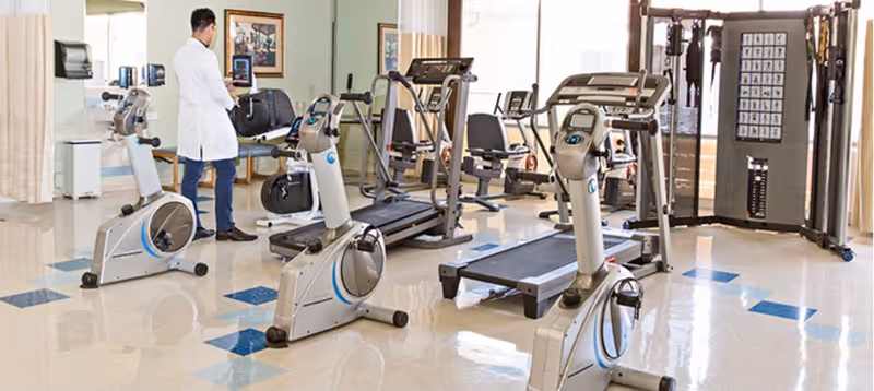 A bright indoor fitness room with exercise bikes, treadmills, weight machines, and a staff member in a white coat using a tablet.