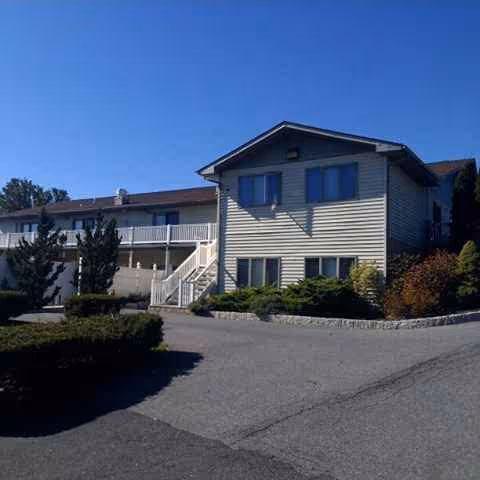 Two-story senior living building with stairs, landscaping, and paved driveway under clear blue sky.