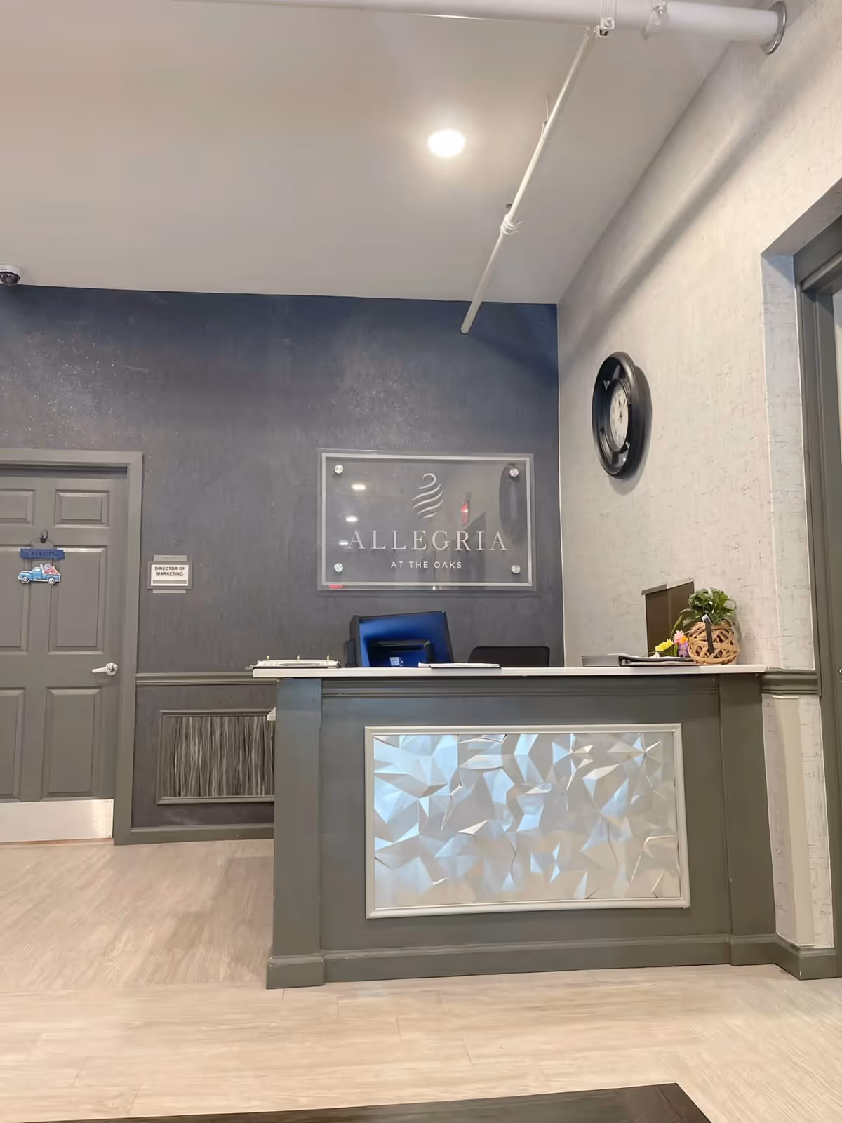 Reception desk area inside Allegria at The Oaks facility with a decorative front panel, a computer monitor, a small plant, and a wall clock. Behind the desk is a dark accent wall with a clear sign displaying the facility name. A door labeled 'Director of Marketing' is visible to the left.