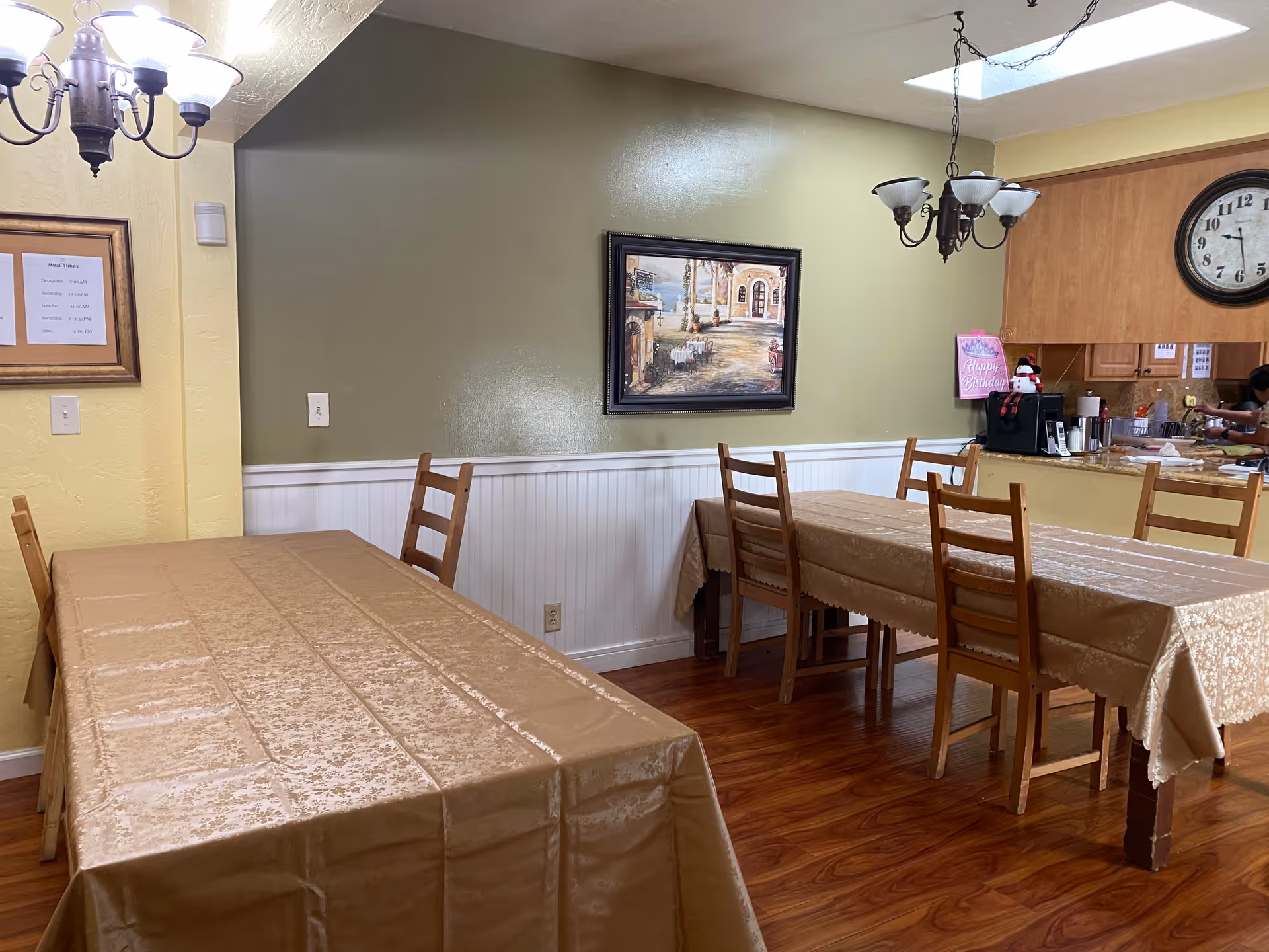 Dining area with two tables covered in beige tablecloths and wooden chairs around them. The room has light green and yellow walls, a framed painting on the wall, a chandelier, a clock, and a kitchen counter in the background with a person partially visible.