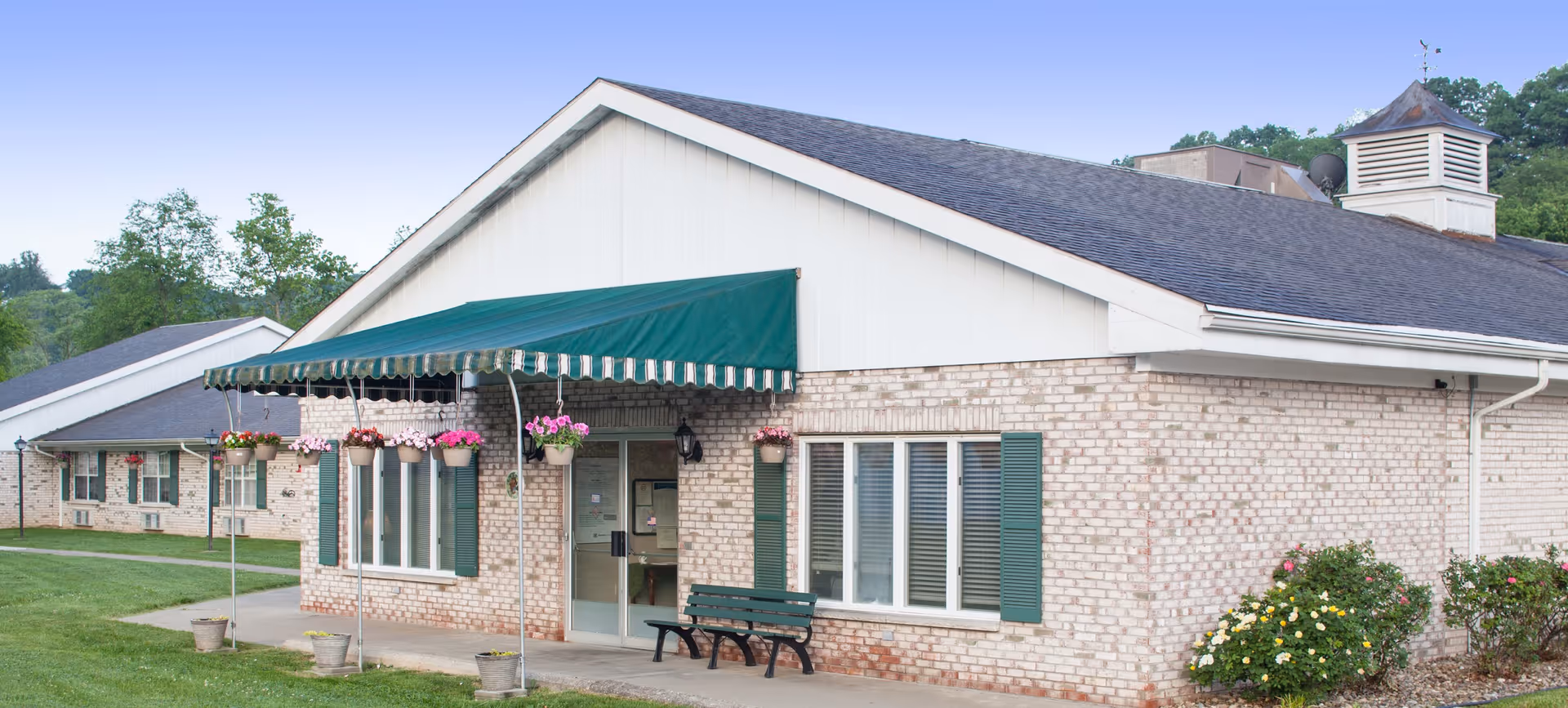 Front exterior of a single-story brick senior care building with a green awning over the entrance, hanging flower baskets, and a bench.
