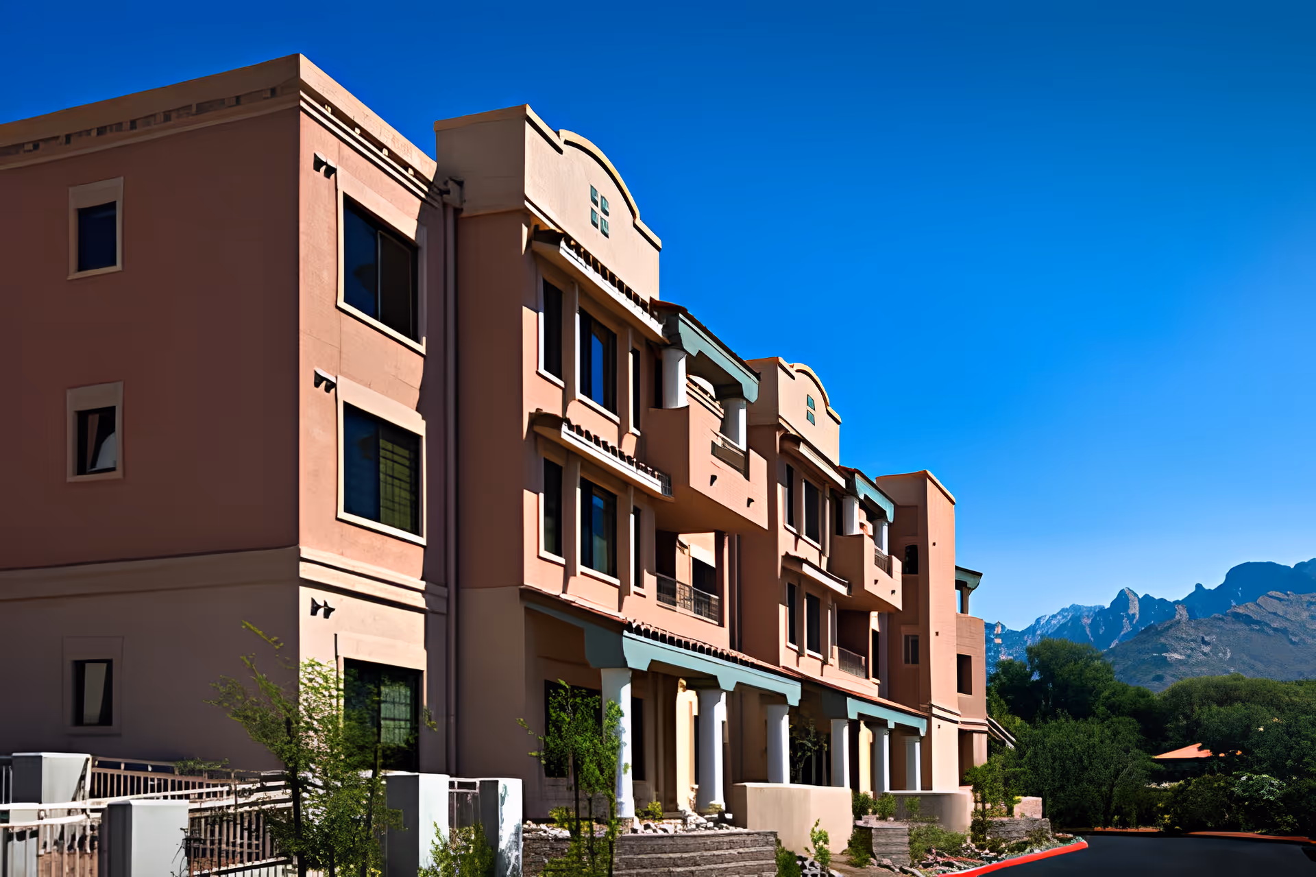 Exterior view of a multi-story senior living facility building with a peach-colored facade, multiple windows, balconies, and white columns at the entrance. The building is set against a clear blue sky with mountains and greenery in the background.