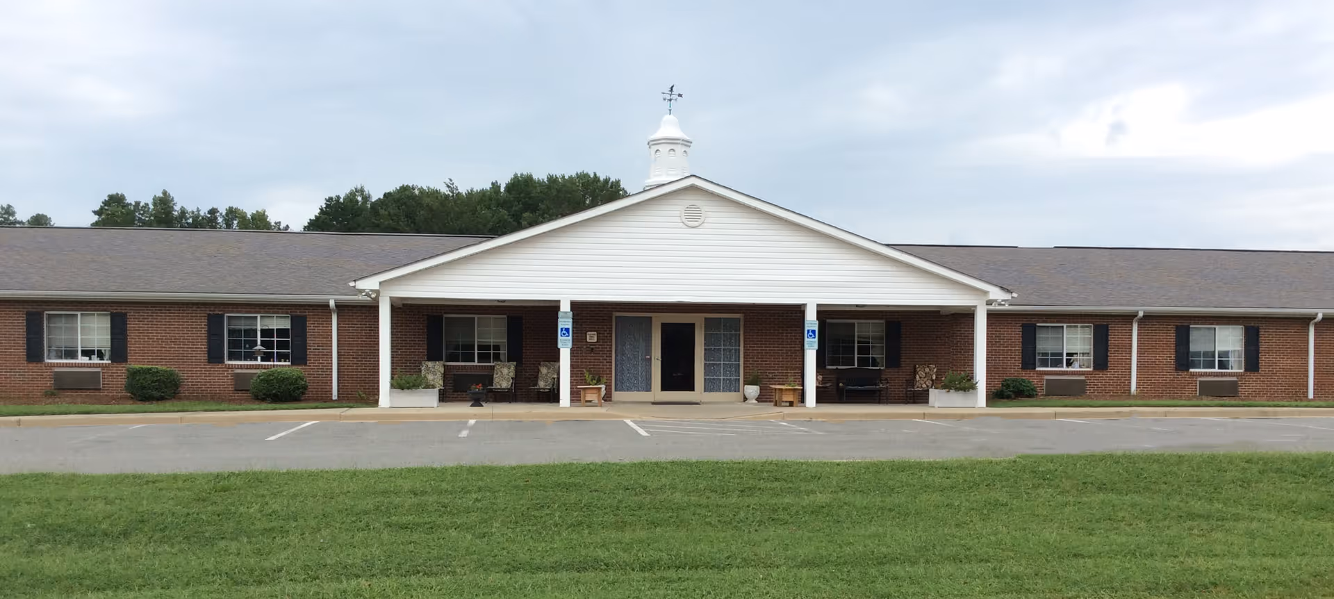Front exterior view of a single-story brick building with a white gabled entrance featuring a small cupola on top. The building has several windows with black shutters and a paved parking area with marked handicap parking spaces in front. There is a grassy lawn in the foreground under a cloudy sky.