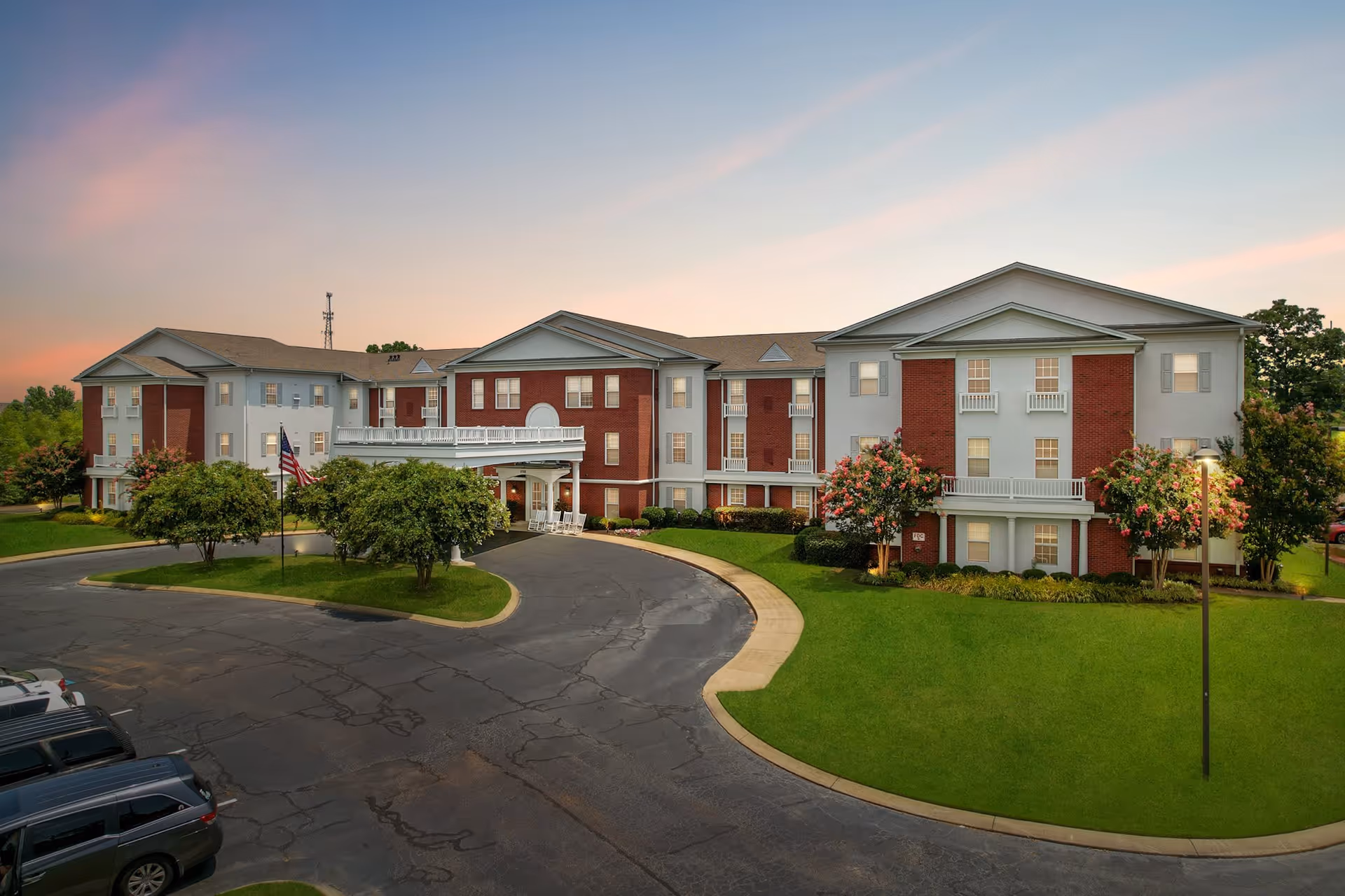 Exterior view of a three-story senior living facility building with a combination of red brick and light gray siding, surrounded by green lawns, trees, and a curved driveway with parked cars under a colorful sunset sky.