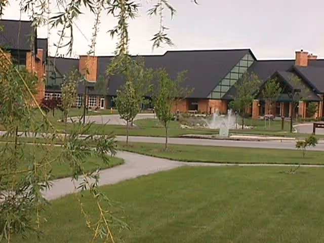 Exterior view of a senior living facility building with a black roof and brick walls, surrounded by green lawns, small trees, and a paved walkway. A water fountain is visible near the entrance.