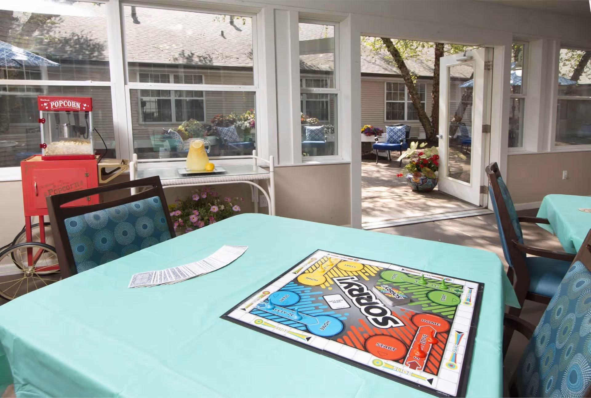 A bright indoor seating area with tables covered in light blue tablecloths. One table has a Sorry board game and a deck of cards laid out. In the background, there is a red popcorn machine and a white cart with a pitcher of lemonade and a lemon. Large windows and a glass door open to an outdoor patio with blue cushioned chairs and potted plants.