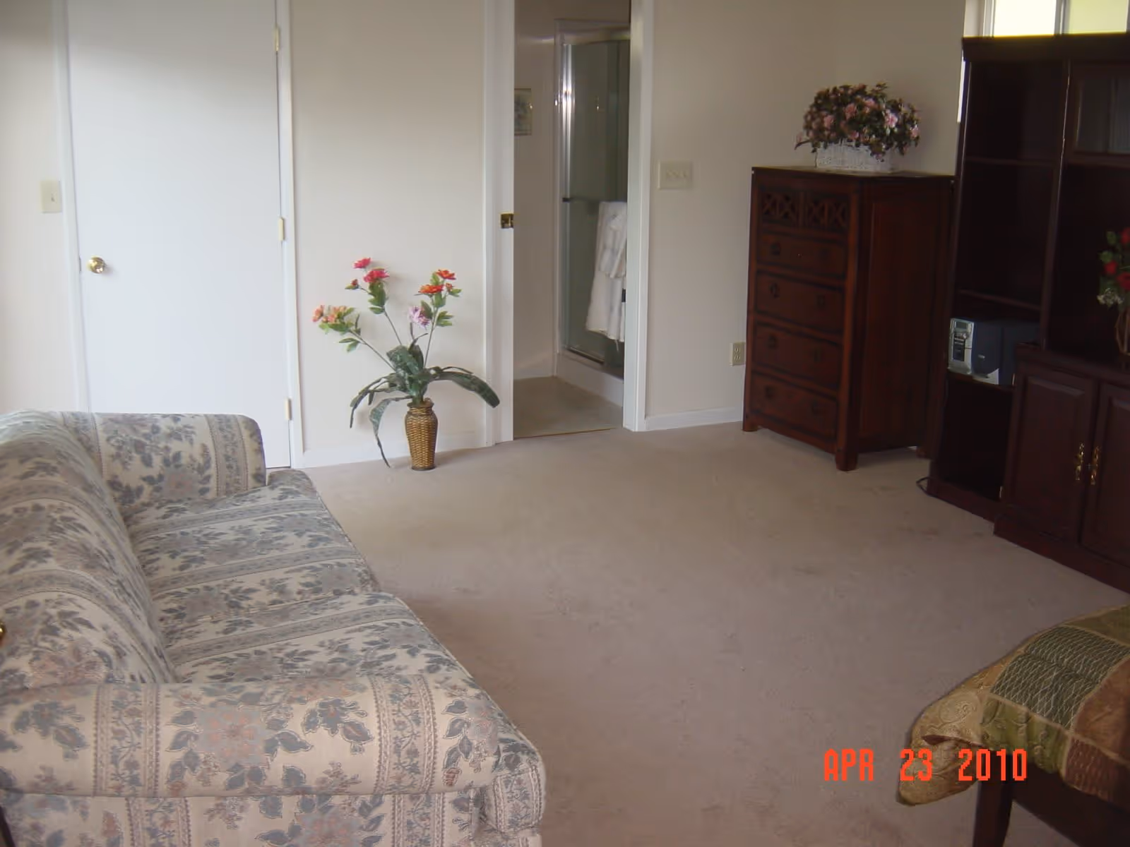 A room with a floral patterned sofa on the left, a vase with artificial flowers on the floor near a white door, a wooden dresser with a flower arrangement on top, and a wooden entertainment center on the right. There is an open door leading to a bathroom with a glass shower door. The carpet is beige and the walls are light-colored.