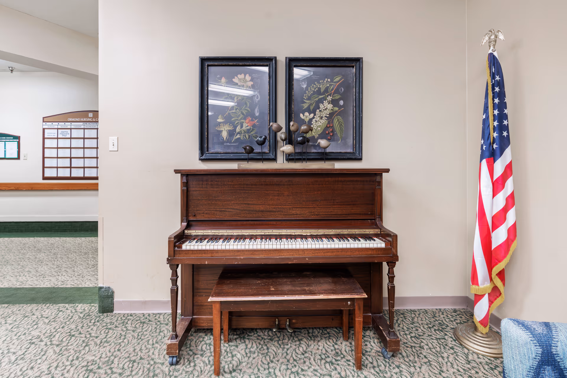 An upright wooden piano with a matching bench in front of it, placed against a beige wall. Above the piano are two framed botanical prints and a decorative piece with bird figurines. To the right of the piano stands an American flag on a gold-colored stand. The floor is carpeted with a patterned design, and a partial view of a blue and white blanket or cushion is visible on the right edge. In the background to the left, a bulletin board with the title 'Ormond Nursing & Care Center' is mounted on the wall.