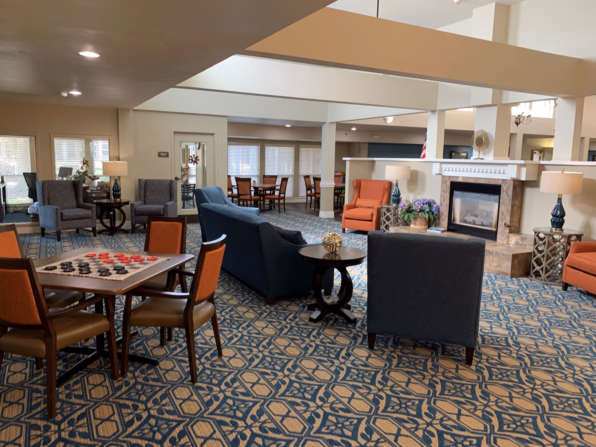 Spacious senior living lounge with patterned carpet, seating, a fireplace, and a game table.